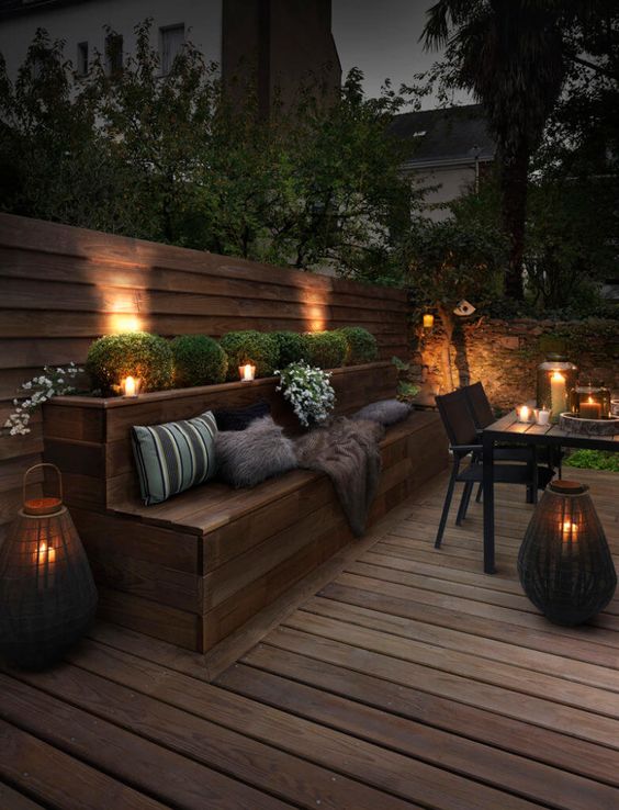 a moody outdoor space clad with wood, with a built in wooden bench and wooden dining set, greenery, built in lights and lanterns