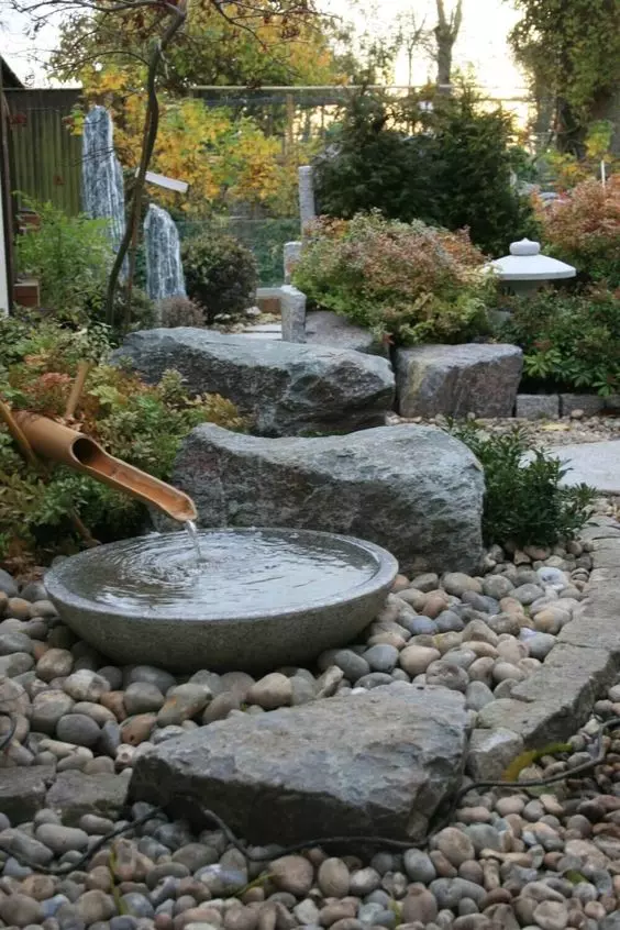 a Japanese front yard garden with a traditional bamboo fountain, pebbles and large rocks, greenery and a candle lantern