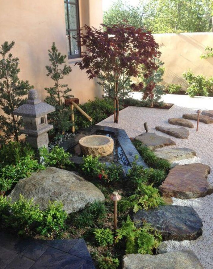 a Japanese front yard garden with a traditional stone lantern, some trees, a traditional fountain of bamboo and a stone bowl plus large rocks