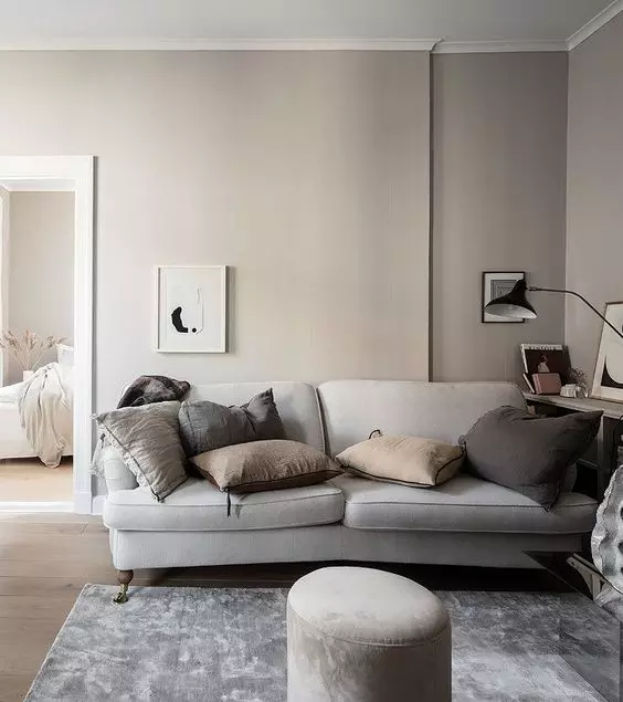 a Nordic living room with greige walls, a grey sofa and dark pillows, a grey rug and a pouf plus a black lamp