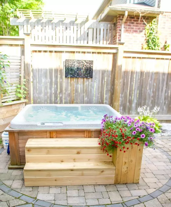 a backyard with a stone floor and a wooden clad hot tub, with wooden steps and a wooden planter with bright blooms