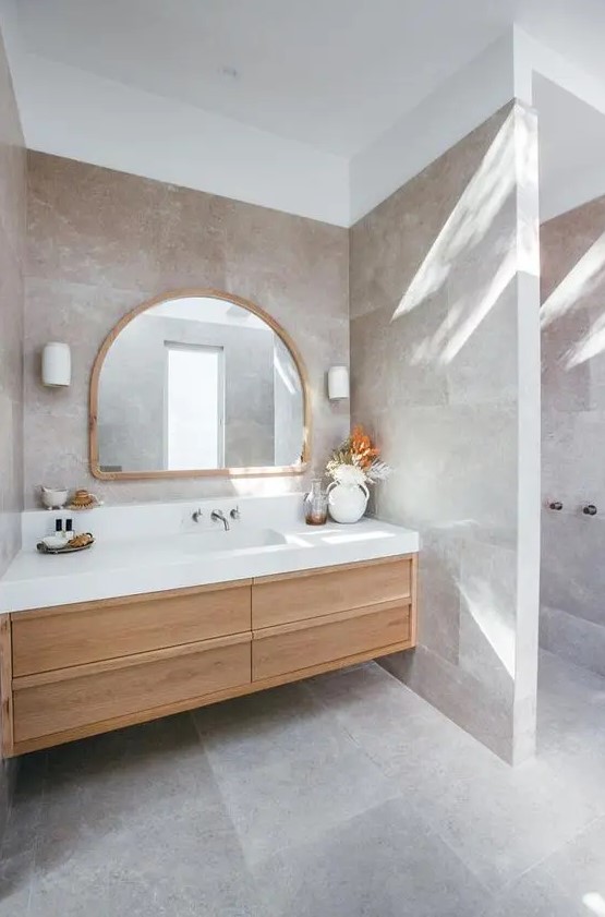a beautiful greige bathroom clad with large scale tiles, a light-stained built-in vanity, an arched mirror and a dried leaf arrangement