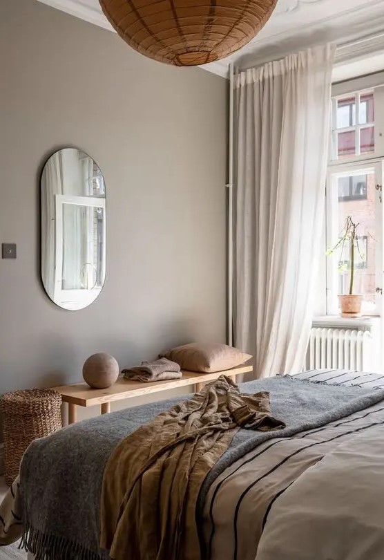 a beautiful greige bedroom with a bed with neutral and printed bedding, a wooden bench, warm-colored textiles, a paper pendant lamp and a basket for storage