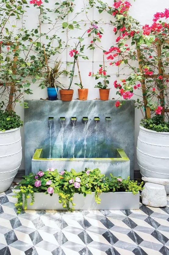 a beautiful modern fountain of a wall with multiple faucets, a concrete tub and a matching planter with blooms next to it