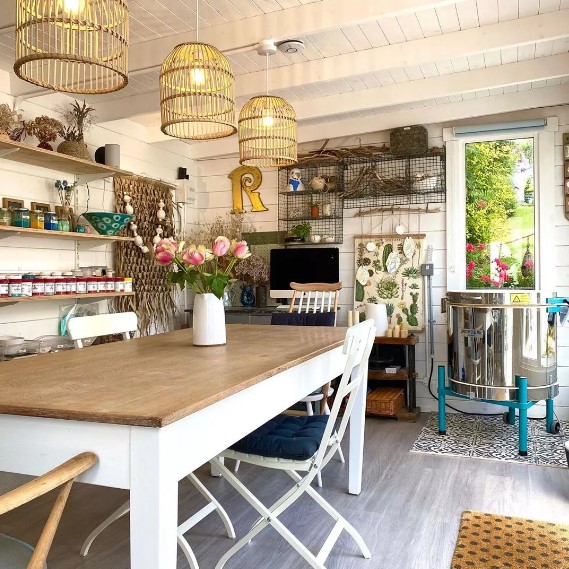 a boho farmhouse she shed with a dining table and chairs, open shelves, a TV, some wire shelves and cage pendant lamps