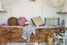 a boho she shed space with a built-in bench that is a chest, pillows and cushions, a canopy with lights, a small crate table with lanterns