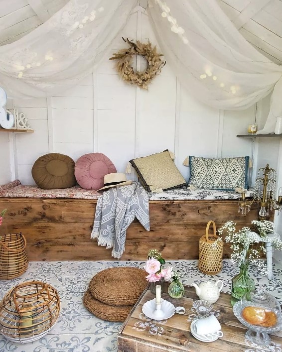 a boho she shed space with a built in bench that is a chest, pillows and cushions, a canopy with lights, a small crate table with lanterns