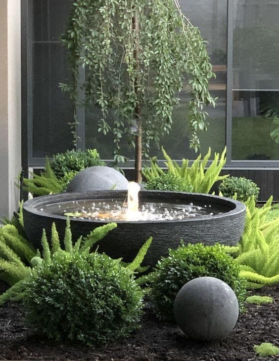 a bold small modern garden with lots of greenery, a large bowl fountain and matching black spheres to add an edgy touch