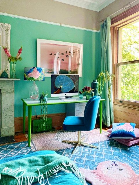 a bright home office with emerald walls, a fireplace, a green and blue desk, a blue chair, pink and blue textiles and a pink artwork