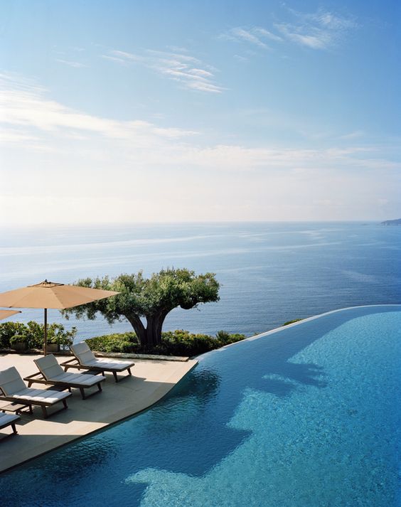 a catchy infinity pool with a sea view, a small terrace with loungers and a parasol and greenery and a tree next to the pool