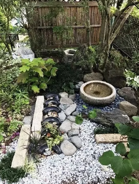 a chic Japanese front yard with pebbles, rocks, a stone bowl with a fountain, trees, greenery and bamboo