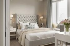 a chic greige bedroom with a grey upholstered bed, neutral bedding, a white vanity and a grey chair, blue curtains and some elegant lamps