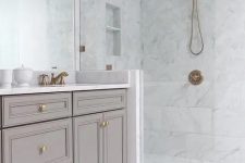 a chic white bathroom clad with marble tiles, with a greige vanity, brass and gold fixtures and a large mirror that takes a whole wall