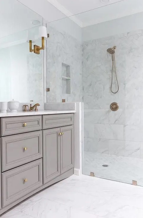 a chic white bathroom clad with marble tiles, with a greige vanity, brass and gold fixtures and a large mirror that takes a whole wall