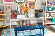a colorful and fun home office with a bright gallery wall, a white desk, a blue polka dot bench, colorful books and blankets