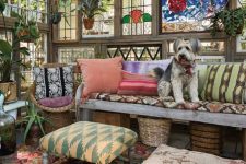 a colorful boho she shed with stained glass, a reclaimed wood sofa, stools and poufs, pillows and potted greenery