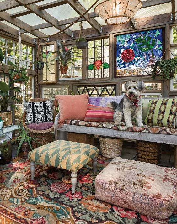 a colorful boho she shed with stained glass, a reclaimed wood sofa, stools and poufs, pillows and potted greenery