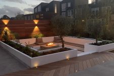 a contemporary sunken fire pit with a built-in bench with taupe upholstery and pillows and a fire pit plus greenery all around