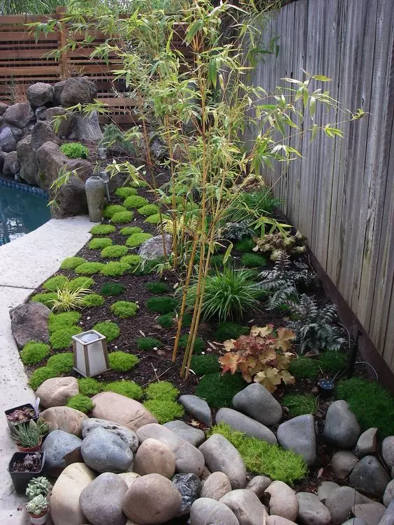 a cool Japanese garden with greenery and leaves, rocks and pebbles, a small tree and some candle lanterns
