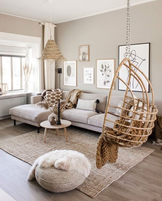 a cool greige living room with a beige sofa with pillows and blankets, a rattan suspended chair, a jute pouf, a woven pendant lamp