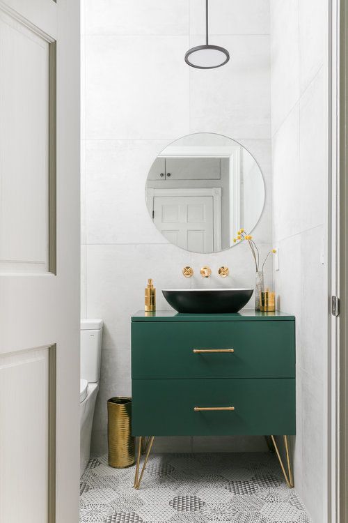 a cool modern powder room with white large scale tiles, a printed tile floor, a green vanity with a black bowl sink, gilded touches for more chic