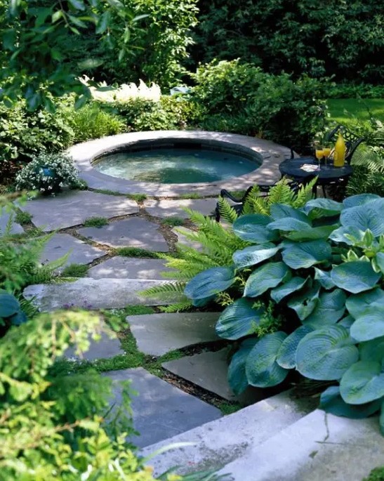 a cute hot tub space right in the garden, with a stone path, a built in tub clad with stone, a couple of chairs for placing drinks