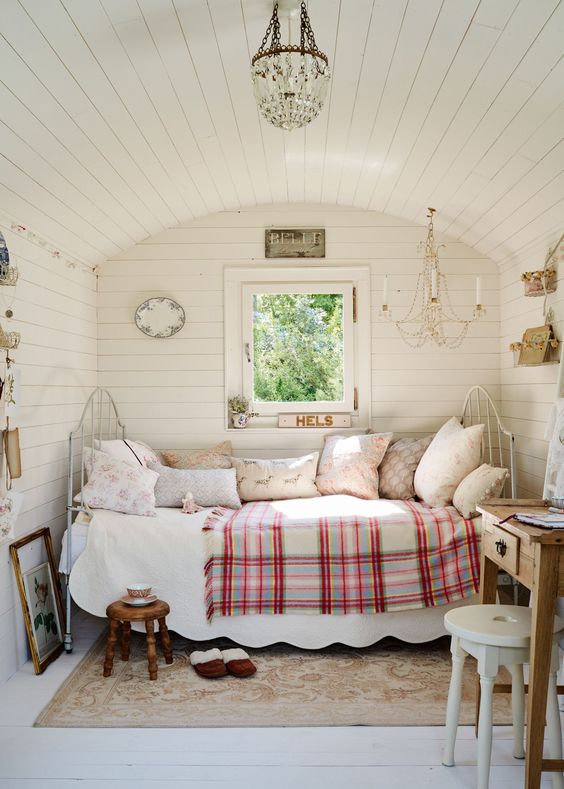 a cute neutral vintage she shed with a metal bed with lots of pillows, a stained desk, stools, a chandelier and some artwork