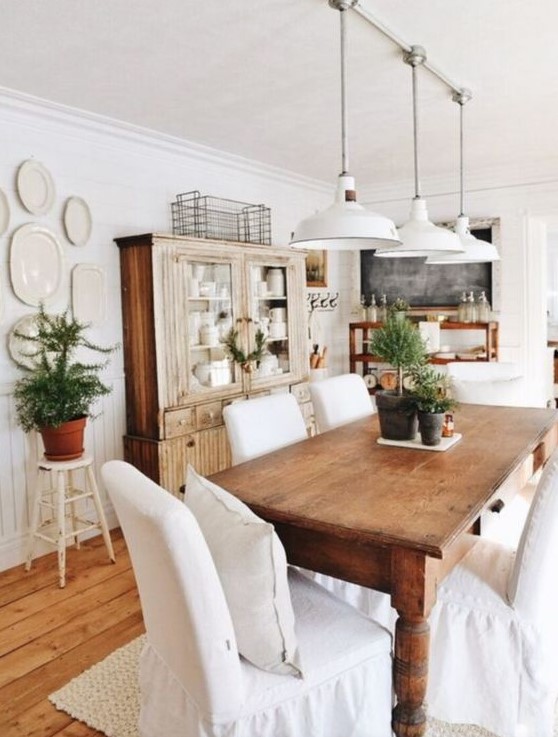 a farmhouse dining area with stained furniture, white covered chairs, potted greenery and white pendant lamps