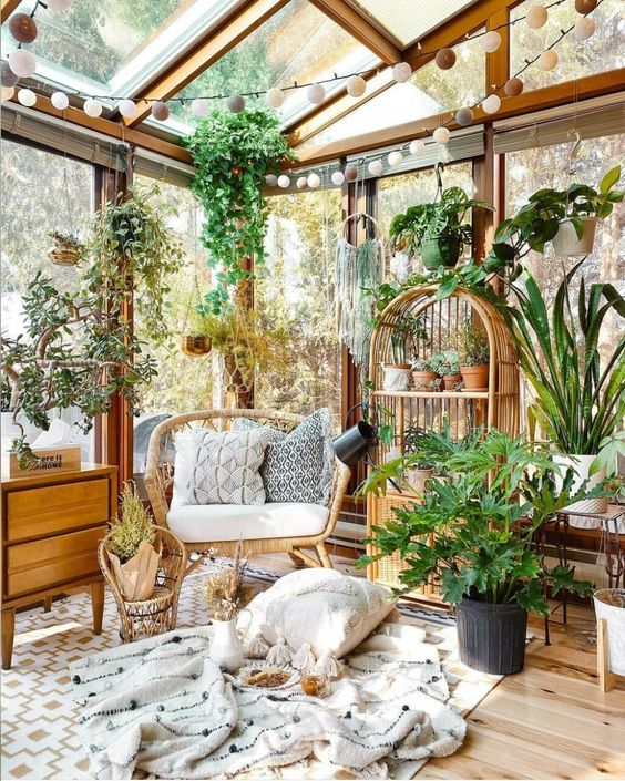 a former greenhouse turned into a she shed in boho style, with wood and rattan furniture, lots of potted plants, pillows and blankets