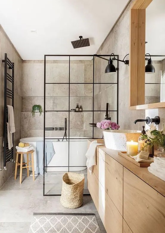 a greige bathroom clad with concrete, with a light-stained vanity and a large mirror, a shower space with a black space divider and black fixtures