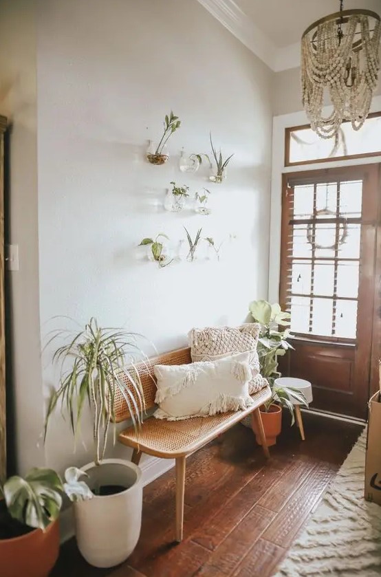 a greige boho entryway with a cane bench, potted plants, a wooden bead chandelier, wall mounted clear planters with greenery