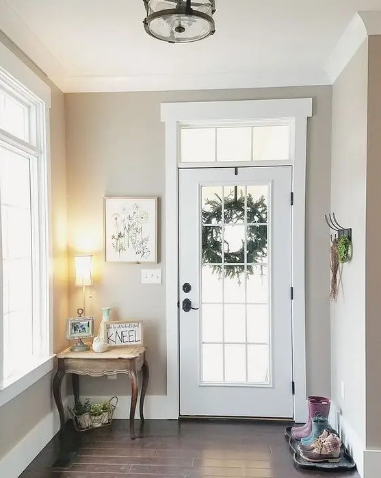 a greige entryway with lots of natural light, a vintage console table, a lamp, some greenery, an artwork and a rack