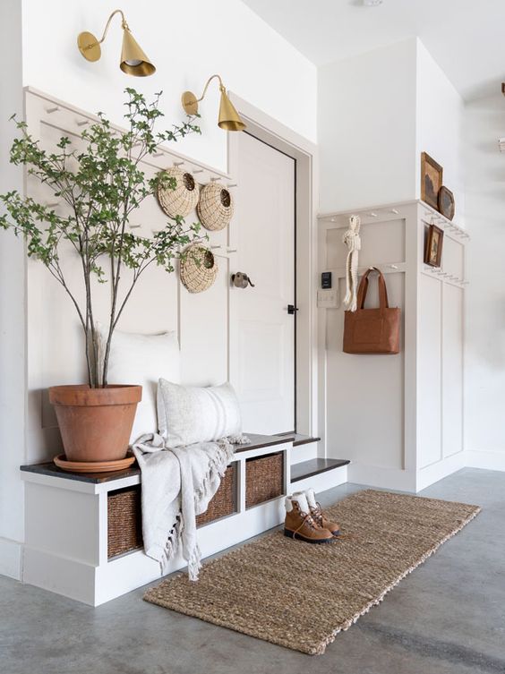 a greige entryway with paneling, a built in storage bench, a rug and a potted plant plus elegant brass sconces