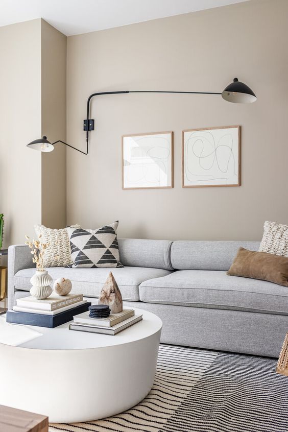 a greige living room with a grey sofa and printed pillows, a round white coffee table and a cool black sconce for an accent