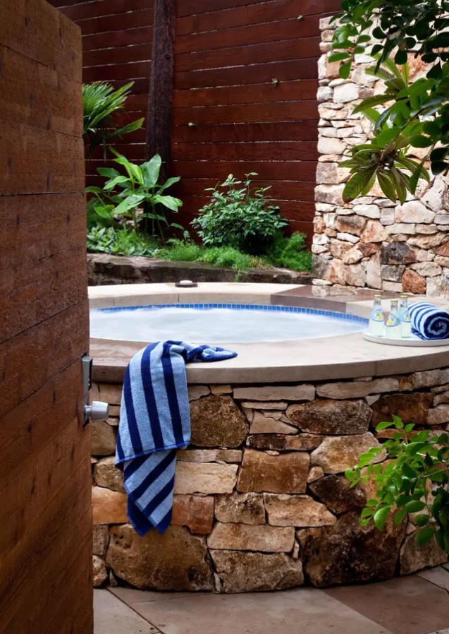 a hot tub nook with a wooden screen, a stone wall, a tub clad with the same stone and some greenery around