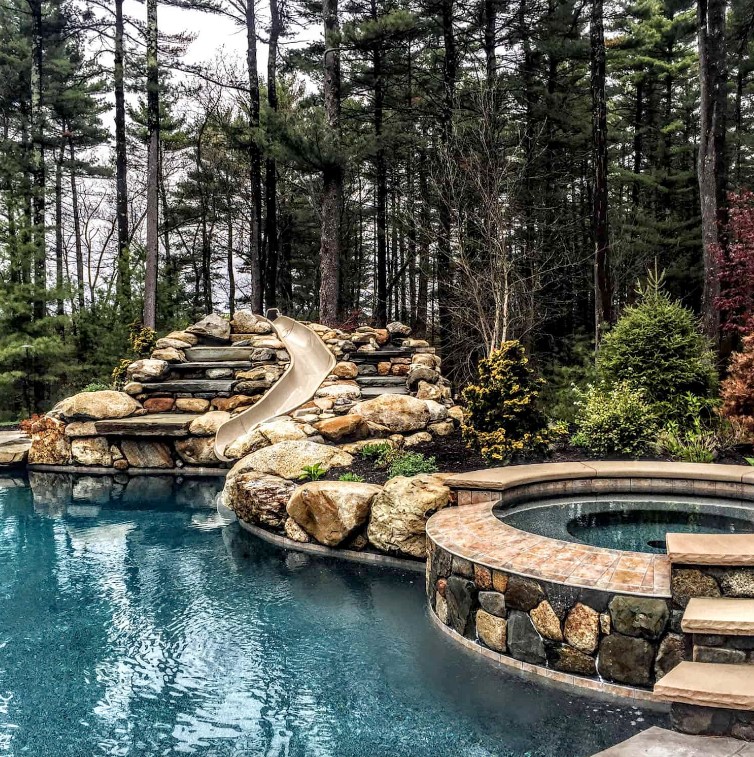 a large pool with a slide and a hot tub all clad with stone and stone tiles plus forest around make the space look ultimate