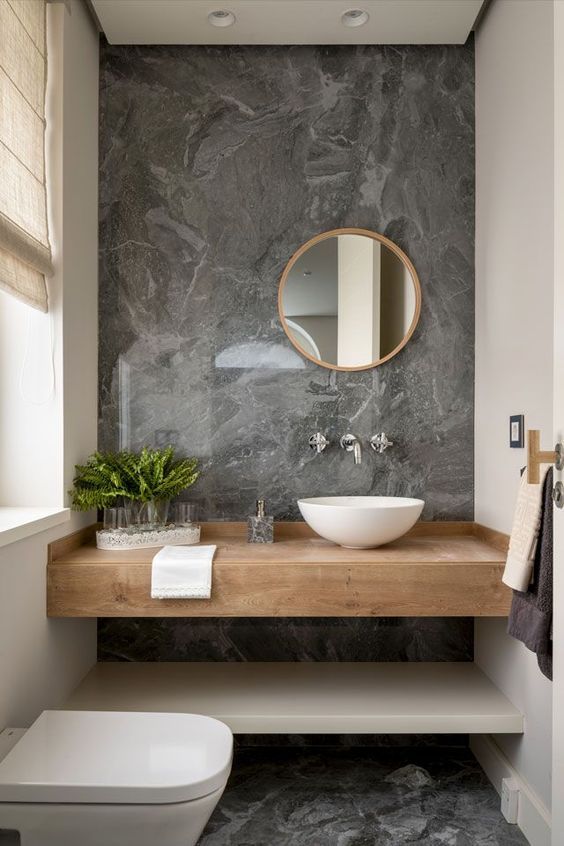 a minimalist powder room with grey stone tiles, a floating vanity, a round mirror, greenery and a neutral toilet