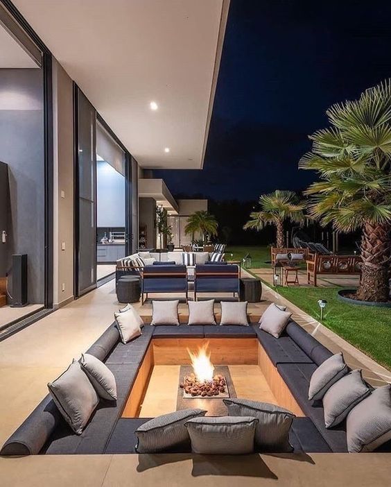 a minimalist sunken patio with built-in benches and grey upholstery and pillows, a fireplace and greenery around