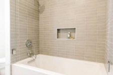 a modern bathroom with greige skinny tiles, a white tub and a niche shelf is a stylish bathroom space