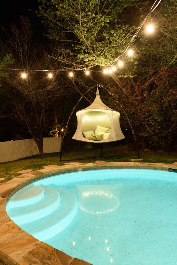 a modern round pool with a stone deck around it, with built-in lights and a tripod pendant chair