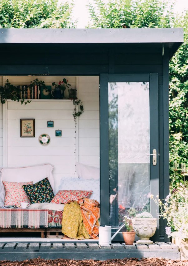 a modern she shed with a glass door, a crate bed with lots of colorful pillows, an open shelf with books and plants
