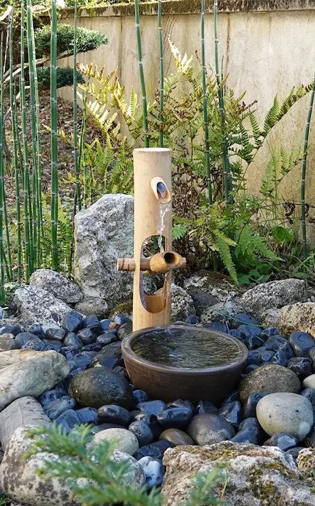 a modern take on a traditional Japanese bamboo fountain with a stone bowl and rocks around plus greenery