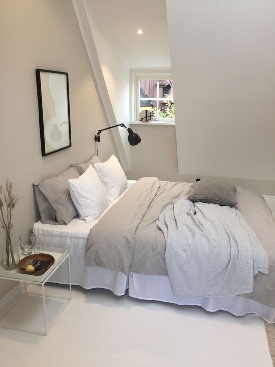 a pretty attic greige bedroom with a small window, a bed with neutral bedding, a nightstand and a black sconce