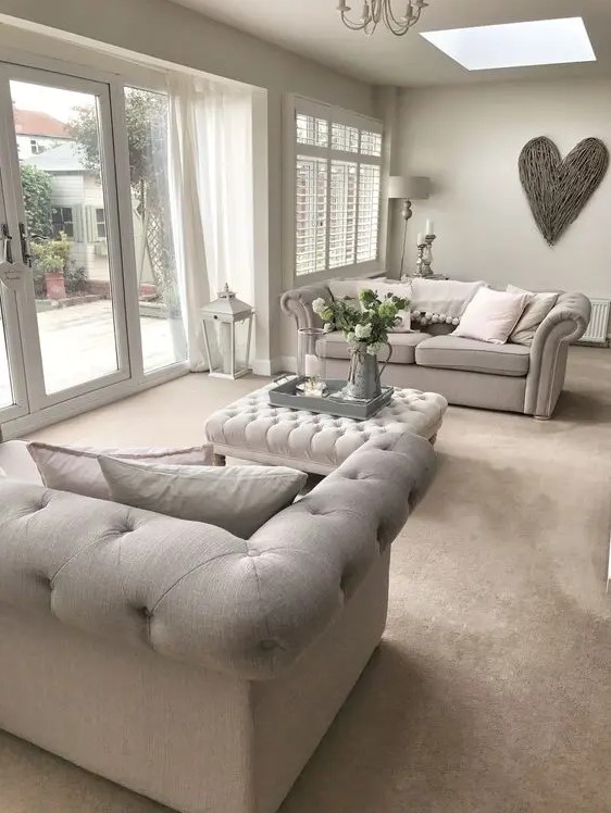 a pretty greige living room with matching tufted sofas, a tufted ottoman, a chandelier, a driftwood heart and some elegant floor and table lamps