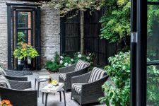 a pretty townhouse courtyard clad with stone tiles, with dark wicker furniture, side tables, greenery and blooms