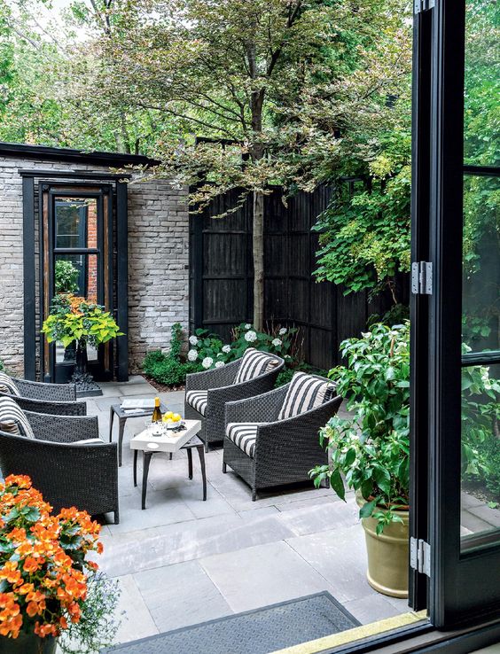 a pretty townhouse courtyard clad with stone tiles, with dark wicker furniture, side tables, greenery and blooms