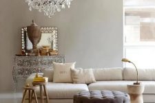 a refined greige living room with a neutral sectional, a taupe tufted pouf, side tables, a crystal chandelier and a beautiful ornated sideboard is wow