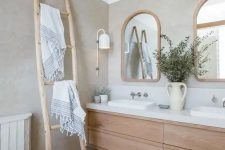 a serene and welcoming greige bathroom with a light-stained floating vanity, a ladder, arched windows in wooden frames and a cool sconce