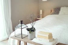 a serene greige bedroom with a bed with neutral bedding, a wooden bench and pendant lamps, some books and plants