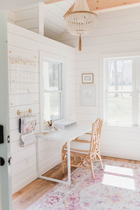 a she shed home office with white planked walls, a wooden bead chandelier, a white desk and a rattan chair, a prink printed rug